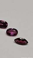 Three dark pink-purple rhodolite garnet gemstones, two oval cuts and one marquis cut, laid on a neutral background displaying facets and color.