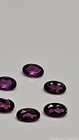 Six oval cut deep fuchsia pink-purple lotus garnet gemstones arranged on a grey background, showing facets and reflections consistent with high clarity.