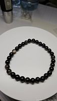 Full view of black round silver sheen obsidian beads arranged in a circular stretchy bracelet, displayed on a white round surface.