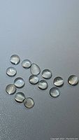 Eighteen small translucent white moonstone cabochons on a flat grey surface showing smooth polished rounded tops and natural adularescent flash.