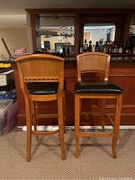 Photo showing front and back views of the two wooden bar stools with black vinyl seats placed in front of a home bar.