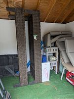 Photo showing two woven chaise loungers standing upright against a wall inside a shed. Loungers are brown woven material with frame and mesh backing visible.