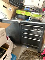 Black Craftsman metal tool storage cabinet with four drawers and one side storage door, placed in garage surrounded by boxes and bags.