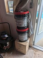 Two stacked black pails with red lids labeled Select Driveway Filler & Sealer, placed on top of a cardboard box next to a vacuum cleaner and near a door.