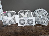 Four fans side-by-side on floor: two square Lasko box fans, one round Lasko Cyclone fan, one rectangular Bionaire window exhaust fan.