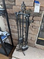 Fireplace tool set displayed standing on floor against a stone wall with metal shelving nearby. Shows the entire 5 piece set on stand with spiral twisted handle top.