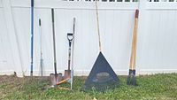 Garden tools lined up against a white fence including shovels, plastic rake, post digger, and metal rake