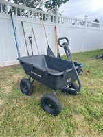 Black Gorilla Carts poly dump cart outdoors on grass near white fence, with garden tools in background.