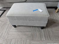 Front view of beige West Elm ottoman on carpeted floor showing upholstery and wooden legs.