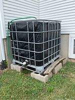 Photo showing the front and side view of the rain barrel in the metal cage with green hose attached and cinder block base.
