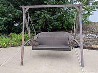 Full front view of the outdoor woven swing seat suspended from the metal frame showing woven seat, armrests, and frame with rust spots.