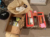 Photo of multiple boxed model train cars including Lionel locomotives and freight cars, cards inside a cardboard box, and a plastic bag with some contents.