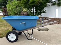 Side view of the blue plastic wheelbarrow showing wooden handles, single black rubber wheel with white rim, and metal support frame.