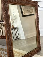 Full view of the antique mirror standing vertically, showing the elaborate carved wooden frame with scroll and floral resin decoration. Reflection shows a room interior.