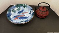 Photo showing ceramic bowl with multicolor fish design beside red and black ornate metal teapot with handle