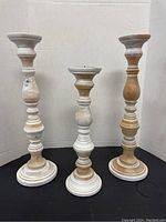 Set of three tall wooden candlesticks in natural and white painted finish with turned spindle work, staged against neutral background on black surface