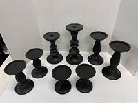 Front view of 9 black candlesticks of various heights and pedestal designs shown against white backdrop.