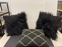 Two black fuzzy pillows and one black pillow with gold decorative pattern arranged on a black surface against a white background.