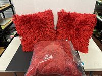 Two red fuzzy pillows and one red pillow in clear plastic bag displayed on a table with plain white background.