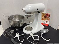 White Kitchenaid Ultra Power Plus stand mixer with bowl, three attachments and recipe book on a black surface and white background.