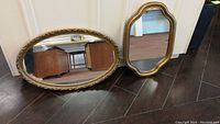 Both oval mirrors resting on dark wood floor against white wall, showing overall style and size difference.