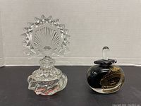 Front view of two perfume bottles with their stoppers in place, one clear fan-shaped design and one black and gold oval shape.