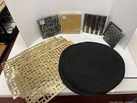 Four gold-colored rectangular placemats, four black round placemats, and five packages of gold and black paper napkins arranged and photographed on a white surface.