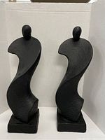 Front view of the two black composite abstract statues side by side showing curved flowing forms and matte textured surfaces on square bases.
