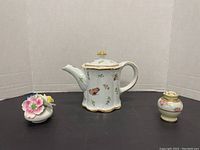 Three porcelain items displayed against a white backdrop: teapot with butterfly and floral decors, small flower figurine, and floral-decorated shaker.