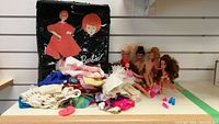 Photo showing six vintage Barbie dolls with different hair styles and colors, multiple pieces of various Barbie clothing, pink and blue shoes, and a black Barbie carrying case with red-haired Barbie illustration.