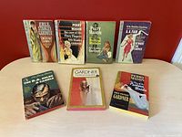 Front view of seven pulp fiction paperback novels showing colorful illustrated covers and titles from the 1950s/1960s, arranged standing on a table.