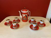Front view of teapot centered with six teacups and saucers arranged around on the table against red background.