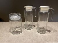 Set of two glass pitchers with white lids and handles, and one glass canister with clear hexagonal lid.