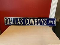Front view of the metal Dallas Cowboys Ave sign on a red background, showing full width of the sign with text clearly visible.
