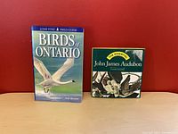 Front view of two bird books placed upright against a red background: 'Birds of Ontario' by Andy Bremer and 'The Essential John James Audubon' hardcover edition.