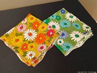 Two retro floral plastic table cloths folded and laying side by side showing one green floral and one yellow floral cloth.