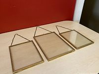 Three brass hanging picture frames laid flat on light-colored table with a red wall background. Two small frames on each side and one larger frame in the middle. Each frame has a glass front and ornate brass border with hanging chain.