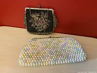 Two vintage clutch handbags on a light wooden surface against a red background: a pastel beaded clam clasp clutch and a floral tapestry purse with a metal kiss lock.