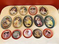 Overview of all 14 Coca-Cola coaster trays arranged on a table showing various vintage lady designs with some rust and wear.