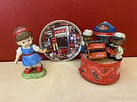 Photo showing three Coca-Cola collectibles on a plain surface with red background: girl figurine, music box carousel with bears, and collectible plate.