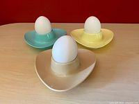 Three pastel color vintage plastic melamine egg cups holding white display eggs, on wooden surface with red background.