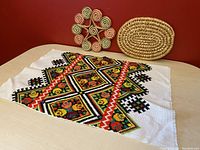 Two woven straw trivets and one Ukrainian needlepoint placemat with a colorful geometric floral pattern on a white fabric background.