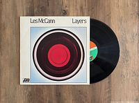 Photo showing the Les McCann 'Layers' vinyl LP partially out of its album jacket on a wooden surface, showing the album front cover design and vinyl label.
