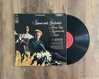 Vinyl LP record visible half out of original album jacket, jacket front cover shows photo of Simon & Garfunkel with floral foreground and dark background, red Columbia record label visible on vinyl.
