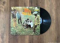 Photo showing the album cover and the black vinyl record partially pulled out to the right, set on a wooden floor. Clear view of the album artwork and record surface.