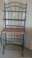 Front view of the wrought iron bakers rack showing dark hunter green frame, wooden middle shelf in honey oak, and decorative scrollwork.