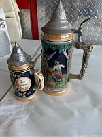 Front view of the two vintage German beer steins showing the detailed painted scenes and pewter lids