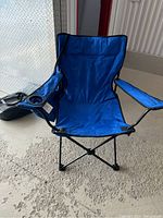 Blue folding camping chair with built-in cup holder on right armrest, shown from front and side angles on rough concrete surface.
