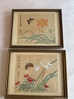 Both framed silk paintings placed side by side on a white surface showing flowers and butterfly designs with Asian script signatures.