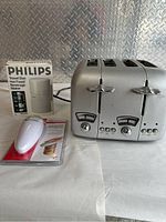 Photo of Philips Travel Duo Hot Food and Beverage Maker in box, a silver 4-slot toaster, and a red and white kitchen tool in its package on white surface.
